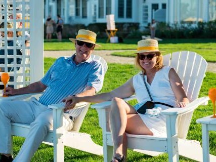 Two guests wearing straw hats relax in white Adirondack chairs on the waterfront lawn at Inn at Perry Cabin, with the resort in the background.